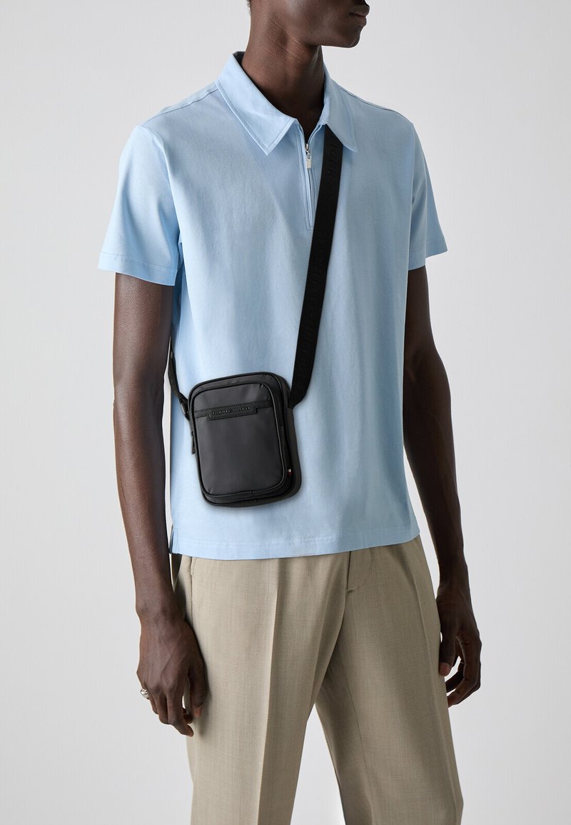 Borsa a tracolla nera in pelle liscia con zip, indossata sopra una polo azzurra. Il modello indossa anche pantaloni beige.