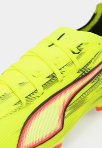 Lys gul fodboldstøvle med sorte detaljer, tekstureret overdel, fire snørehuller og en iøjnefaldende rød og hvid logo på siden.