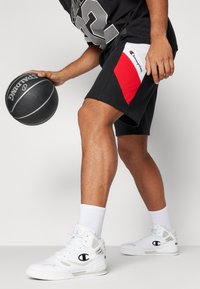 Black basketball shorts with red and white accents, paired with white sneakers and crew socks, holding a black basketball.