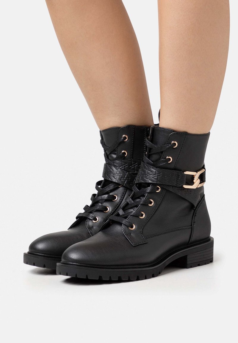 Bottes basses en cuir noir avec semelles épaisses, devant à lacets, et sangles décoratives avec boucles dorées portées sur des jambes nues.