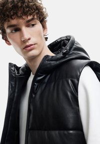 Schwarze, gefütterte Lederweste mit Kapuze und Reißverschluss. Verfügt über gesteppte Abschnitte und eine glatte Textur über einem weißen Kleidungsstück darunter.