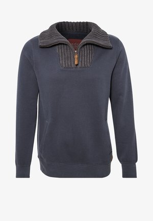 Uomo con la barba che indossa un pullover scuro con collo a zip, jeans blu, stivali marroni con lacci, e porta uno zaino beige su una spalla.