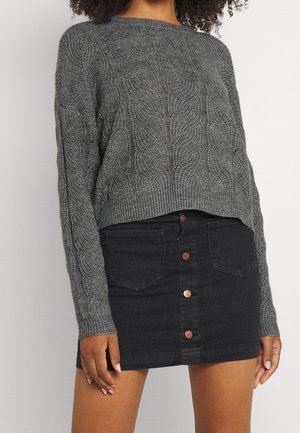 Femme portant un pull gris en maille torsadée et une mini-jupe en denim noire avec boutons sur le devant et détails de poches.