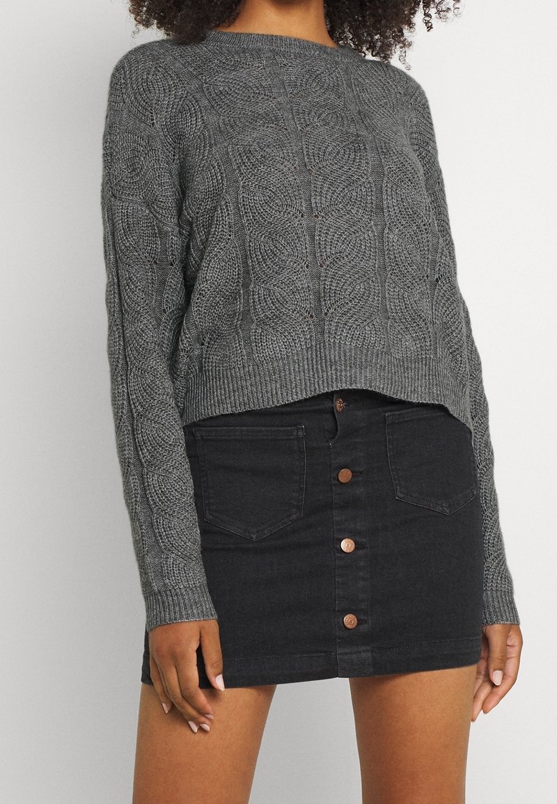 Femme portant un pull gris en maille torsadée et une mini-jupe en denim noire avec boutons sur le devant et détails de poches.