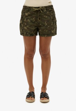 Mujer con pantalones cortos cargo de camuflaje verde con cintura ajustable y sandalias negras sin talón, de pie sobre un fondo blanco.