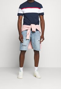 Muž v námořnickém, bílém a červeném pruhovaném tričku, světlých modrých roztrhaných džínových šortkách, růžové mikině uvázané kolem pasu, bílých teniskách a ponožkách.