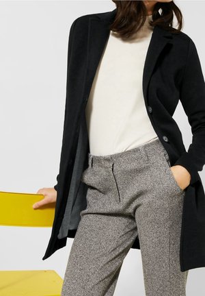 Femme portant un manteau noir, un haut blanc et un pantalon en tweed gris, une main dans la poche et l’autre sur le dossier d’une chaise jaune.