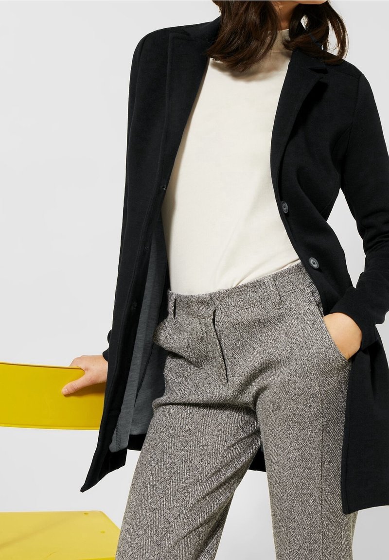 Femme portant un manteau noir, un haut blanc et un pantalon en tweed gris, une main dans la poche et l’autre sur le dossier d’une chaise jaune.