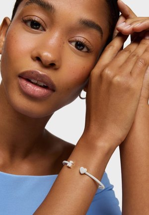 Gros plan sur une femme portant un haut bleu clair et un bracelet en argent avec des breloques en forme de cœur et d'ours en peluche à son poignet.