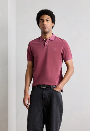 Mand med afro-frisure iført en vinrød polo og sorte jeans, stående med den ene hånd i lommen mod en ensfarvet baggrund.