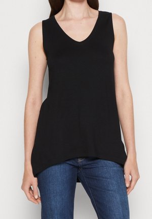 Kobieta w czarnej, bezrękawnikowej bluzce z dekoltem w serek, w niebieskich dżinsach, stojąca na jednolitym jasnym tle.