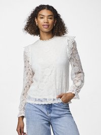 Hvit bluse i blonder med lange, transparente ermer, bølget halslinje og kant, med blomsterdesign og teksturerte detaljer, kombinert med blå jeans.