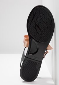 Semelle d'une sandale noire avec un motif texturé, des brides de cheville couleur nude, et un marquage de la marque "Ted Baker London", photographiée sur fond blanc.