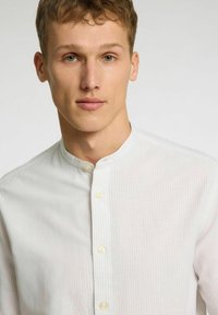 Camisa blanca de botones con cuello alzado, presenta sutiles rayas verticales y una textura suave, hecha de una tela ligera.