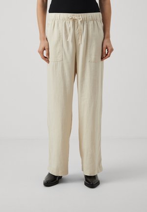 Light beige, loose-fit, drawstring pants with two front pockets and a soft, textured fabric. Paired with black shoes.