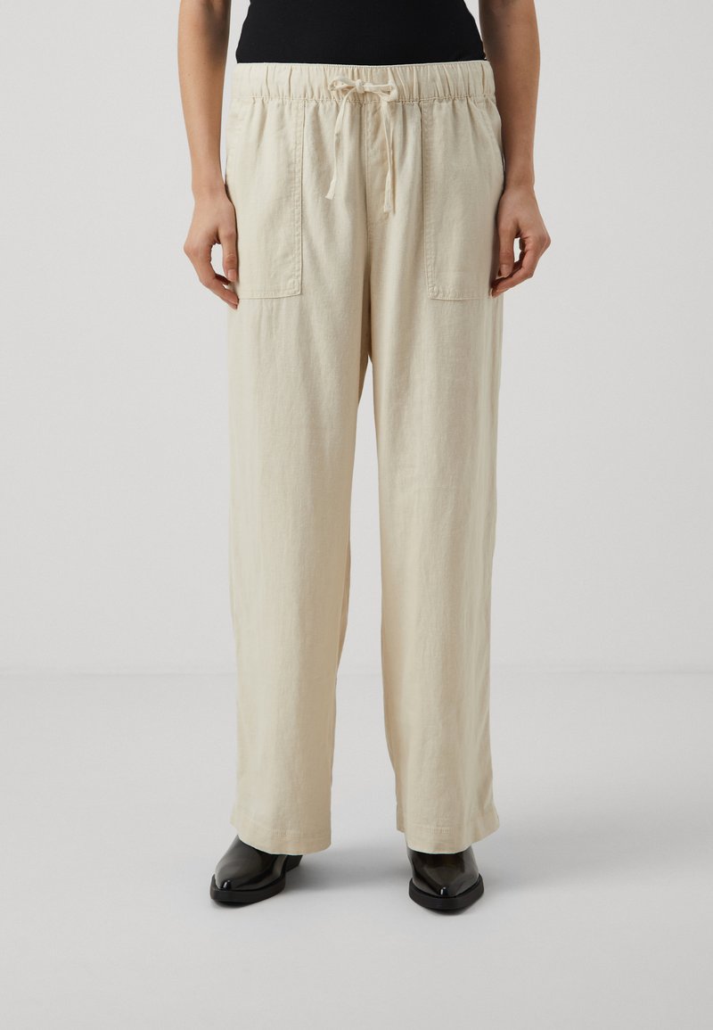 Pantaloni beige chiaro, morbidi e di taglio ampio con coulisse, dotati di due tasche frontali e realizzati in un tessuto morbido e testurizzato. Abbinati a scarpe nere.