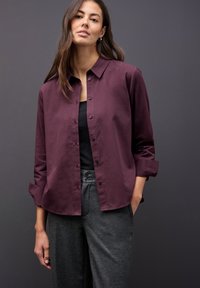 Burgunderfarbendes Button-down-Hemd mit Kentkragen, langen Ärmeln, die an den Manschetten hochgerollt sind, kombiniert mit grauen Hosen, die zwei Knöpfe am Taillenbund haben.