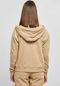 Beige hettegenser med en avslappet passform og stor hette. Myk stofftekstur og ribbestrikkede mansjetter ved håndleddene og kanten. Enkel design uten dekorasjoner.