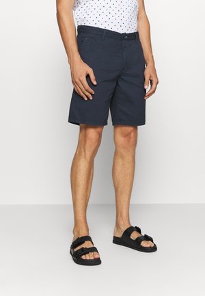Homme portant un short navy, une chemise blanche à motifs et des sandales noires à deux lanières, se tenant devant un fond blanc uni.