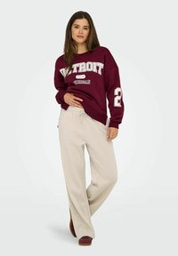 Bordeauxfarbener Sweatshirt mit dem Aufdruck "DETROIT", weißen Akzenten und der Zahl "2", kombiniert mit cremefarbenen weiten Hosen und braunen Schuhen.