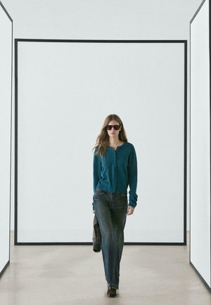 Cardigan lavorato a maglia turchese con dettagli a bottoni, abbinato a jeans di denim scuro. L'ambientazione presenta pareti bianche minimaliste e linee geometriche marcate.