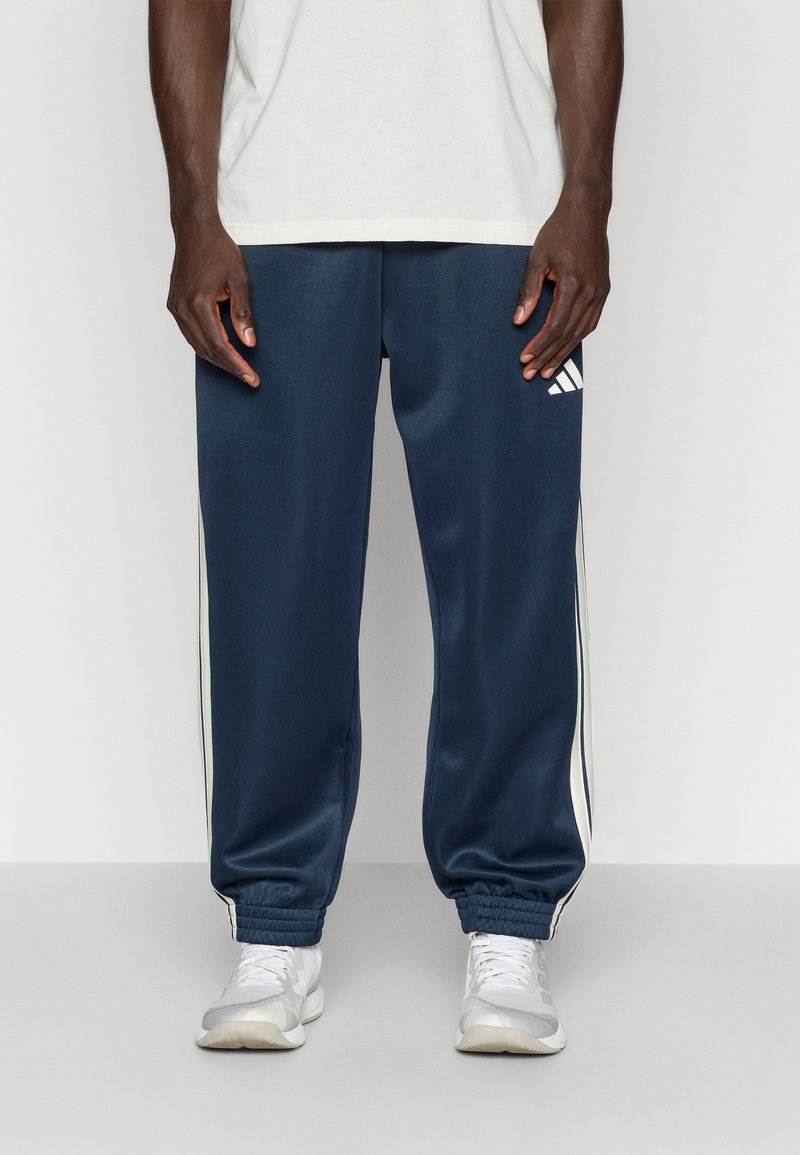 Mornarske trenirke izdelane iz gladkega blaga, z belimi stranskimi črtami in elastičnimi manšetami. Oblikovanje vključuje Adidasovo logo na zgornjem stegnu.