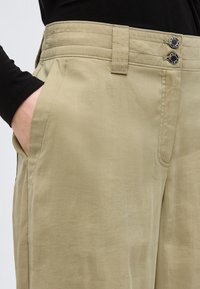 Μπεζ παντελόνι με διπλό μαύρο κουμπί, θηλιές για ζώνη και πλευρική τσέπη, φορεμένο με μαύρο μακρυμάνικο τοπ και χέρι στην τσέπη.