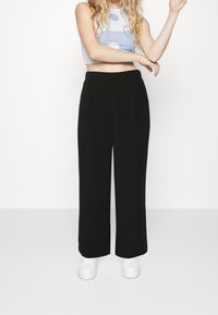 Schwarze, weite Hosen mit Bundfalten, hoher Taille und glatter Textur, kombiniert mit weißen Plateausneakern.