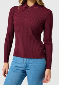 Bordowy prążkowany sweter z kołnierzykiem i półzamkiem. Posiada długie rękawy i falowany brzeg, zestawiony z jasnoniebieskimi dżinsami.