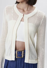 Cárdigan de punto en crema con tejido abierto, cuello redondo y detalles en dorado. Lleva puesto sobre una blusa blanca y combinado con un cinturón azul.