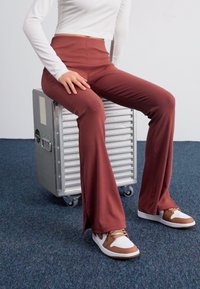 Brune ribbede buks med vidde og høj talje, parret med hvide og brune sneakers. Modellen sidder på en metallisk sølvkasse på et blåt tæppe.