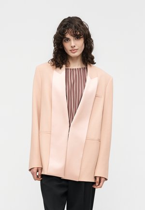 Woman with curly dark hair wears oversized pale pink blazer over brown striped top and black pants, standing against plain background.