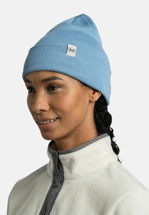 Gorro azul de canalé con parte superior puntiaguda y parche con logo, combinado con una chaqueta de forro polar blanca con ribetes y botones grises.