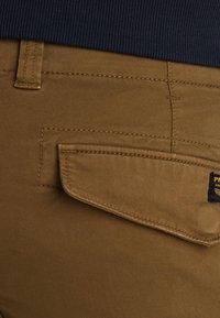 Smeđe pamučne hlače s stražnjim džepom. Džep ima preklopno zatvaranje i žuti logotip. Glatka tekstura s vidljivim detaljima šivanja.
