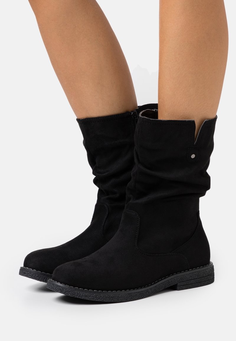 Bottes en daim noir mi-mollet avec bouts ronds, talons en caoutchouc texturé bas et tiges souples portées sur des jambes nues.