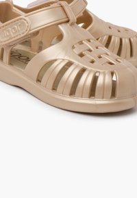 Beige plastic sandals with open cutouts, strap across the instep, and a rounded toe. Smooth texture and flexible design for comfort.