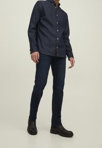 Man som står och bär en mörk denimskjorta med knappar, mörkblå jeans och svarta Chelsea-boots mot en enkel ljus bakgrund.