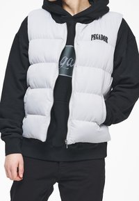 Ljusgrå pufferväst med quiltat mönster, framdragkedja och svart "PEGADOR"-logotyp. Bärs över en svart hoodie med en rund logotyp.