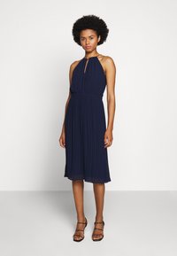 Rochie halter bleumarin cu textură pliată, talie elastică și detaliu cu decupaj la gât, asortată cu sandale cu bretele.