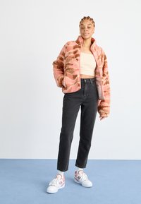 Giacca di pile a motivo rosa e marrone con colletto alto, indossata sopra una maglia beige, abbinata a jeans neri e sneakers multicolori.