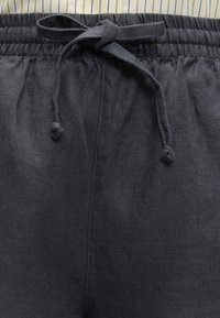 Gros plan de la ceinture d’un pantalon en lin gris foncé avec bande élastique et nœud de cordon noué sur un fond en tissu rayé.