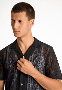 Camisa de manga corta con estampado en negro y gris, con cuello relajado y botones. Textura de punto con diseño de tejido abierto.