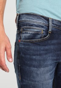 Jeans in denim blu scuro con una texture consumata. Presentano cinque tasche, cuciture gialle a contrasto e un accento rosso vicino alla tasca posteriore.