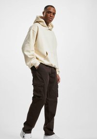 Haut à capuche crème avec une poche avant et un cordon de serrage, associé à un pantalon cargo marron foncé avec des poches latérales. Des baskets blanches complètent le look.