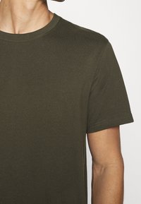 Gros plan d'une personne portant un t-shirt vert olive à manches courtes et col rond sur un fond clair uni.