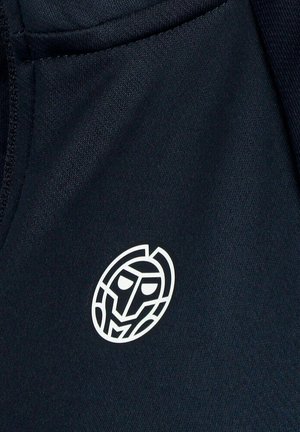 Logotipo abstracto de rostro geométrico blanco sobre tela oscura, centrado cerca del borde de la costura.