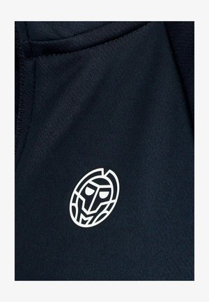 Logotipo abstracto de rostro geométrico blanco sobre tela oscura, centrado cerca del borde de la costura.