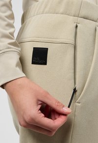Beige polyester broek met textuurstof, voorzien van een zwart logopatch en een verstelbare tailleband met een trekkoord. Close-up van een hand die aanpast.