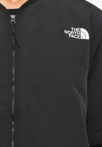 Veste noire zippée avec le logo The North Face blanc sur la poitrine gauche, portée par-dessus un haut noir.