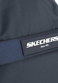 Mochila de tela azul marino con superficie texturizada, que presenta una etiqueta negra prominente con el logotipo blanco "SKECHERS" y el texto "Desde 1992".